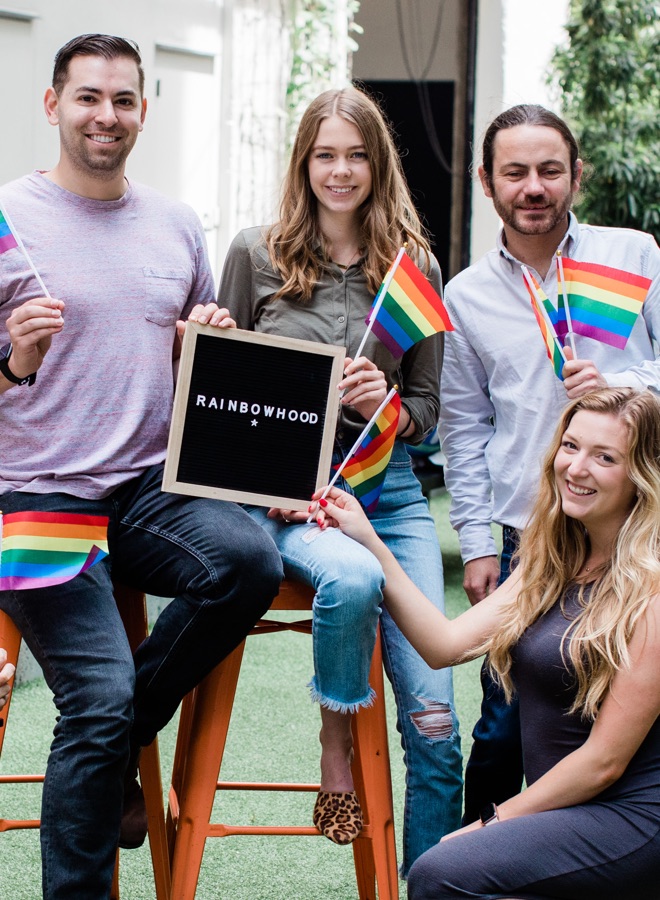 4 teammedlemmer med stolthedsflag, der holder skiltet "Rainbowhood"